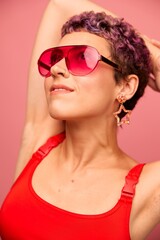 Fashion portrait of a woman with a short haircut in colored sunglasses with unusual accessories with earrings smiling on a pink bright background