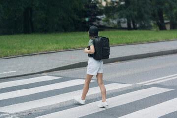 A girl with braids in a black backpack hat crosses the streets at the pedestrian crossing.