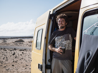 Caucasian man smiling, having a coffee in the morning in his camper van and ejoying the landscape views