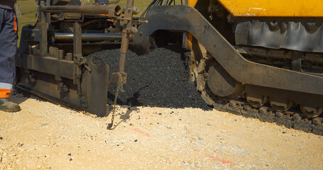 CLOSE UP: Heavy paver machinery dropping fresh black asphalt over crushed rocks