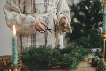 Making Christmas rustic wreath. Woman hands holding fir branches and making wreath on rustic wooden table with ribbon, golden bells, candles. Moody holiday image. Winter holiday workshop
