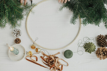 Making modern christmas wreath flat lay. Woman hands with round hoop, ribbon, candle, pine cones,...