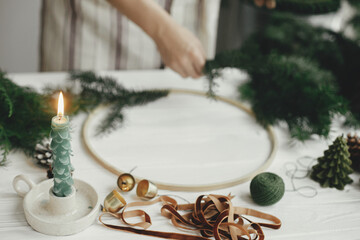 Stylish candle as fir tree, golden bells and ribbon on background of woman hands making boho wreath on white wooden table. Making Christmas modern wreath. Winter decor, moody image