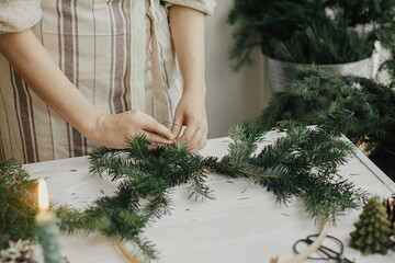 Making Christmas modern wreath. Woman hands with fir branches making wreath on round hoop on white...