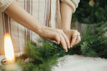 Woman hands holding fir branches and making modern christmas wreath on white wooden table with...