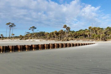 Sunny morning on a wide sandy beach looking toward shore, rusted metal erosion wall, palmetto trees...