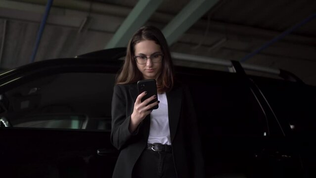 A young businesswoman is standing in the parking lot by the car with a phone. A girl with glasses and a jacket is texting on a smartphone.