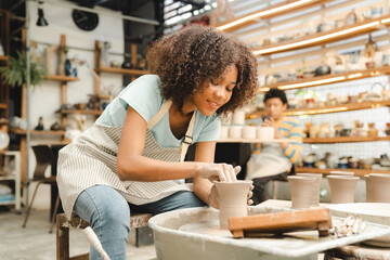 Fototapeta premium Beautiful young woman holding pottery instrument for scraping, smoothing, shaping and sculpting. Lady siting on bench with pottery wheel and making clay pot