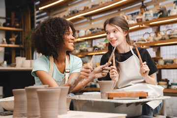 Beautiful young woman holding pottery instrument for scraping, smoothing, shaping and sculpting. Lady siting on bench with pottery wheel and making clay pot