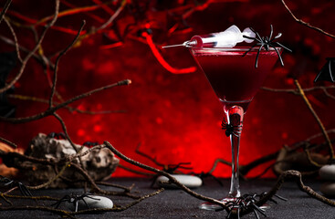 Halloween alcoholic cocktail bloody martini with syringe on scary dark red background with twisted branches, bats, stones, pumpkin guards and spiders, festive drink for vampire party