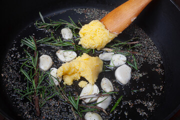 Close-up in a frying pan with olive oil, ghee (clarified butter), garlic cloves, rosemary and herbs. Horizontal top view. Cooking stage.