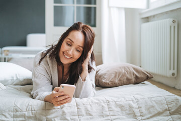 Young woman forty year with brunette long hair in cozy knitted cardigan using mobile phone in bed at home