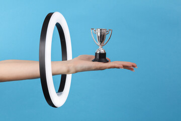 A woman's hand holds a mini silver winner cup through a led ring lamp on a blue background. Creative idea.