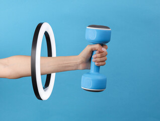 Woman's hand holds plastic dumbbell through led ring lamp on blue background. Creative idea. Fitness, sport concept