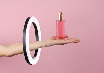 Woman's hand holds perfume bottle through led ring lamp on pink background. Creative idea. Beauty and fashion concept