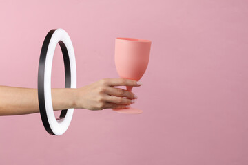 Woman's hand holds plastic glass through led ring lamp on pink background. Creative idea