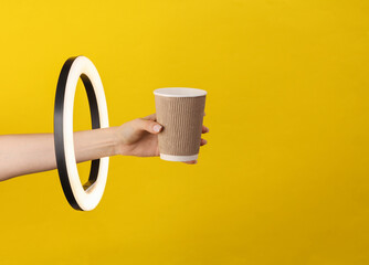 Woman's hand holds paper coffee cup through led ring lamp on yellow background. Creative idea.