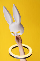 Woman's hand holds white plastic mask from a sex shop through led ring lamp on yellow background....