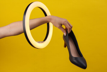 Woman's hand holds heeled shoes through led ring lamp on yellow background. Creative idea. Beauty...