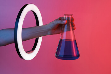 Woman's hand holds chemical laboratory flask through led ring lamp in red blue neon gradient light. Creative idea.