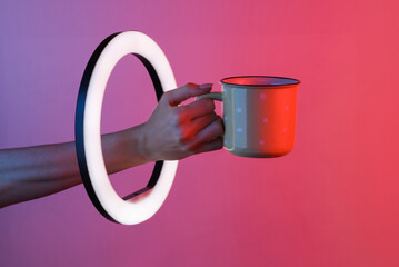 Woman's hand holds ceramic cup through led ring lamp in red blue neon gradient light. Creative idea.