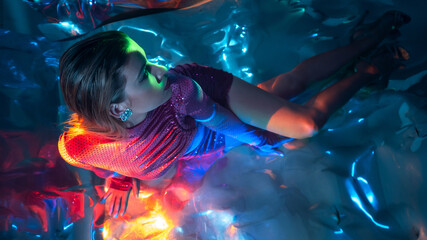 Glamorous portrait of a young beautiful blonde in a stylish scarlet short dress, sitting on the floor, refraction effects of colored light, futuristic fashion shooting in neon light
