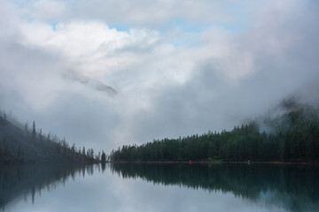 Tranquil meditative misty scenery of glacial lake with reflection of pointy fir tops and clouds at early morning. Graphic EQ of spruce silhouettes on calm alpine lake horizon. Mountain lake in fog.