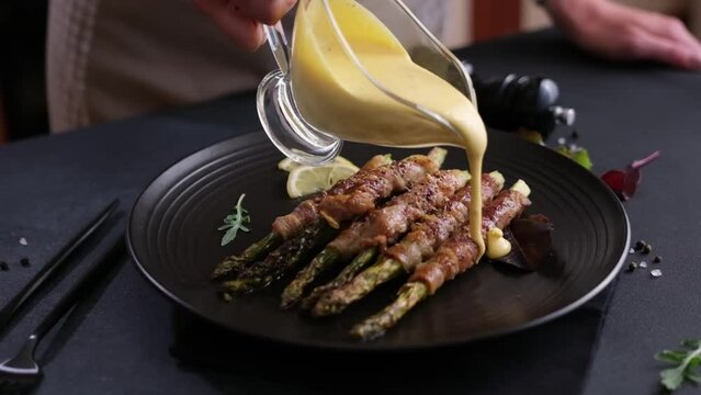 Woman pouring hollandaise sauce onto Baked asparagus wrapped with bacon on a plate