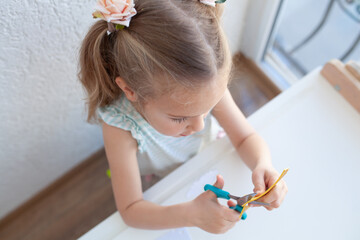 The girl cuts and sculpts from plasticine, is engaged in creativity at a white table against a white wall, early childhood development, kindergarten