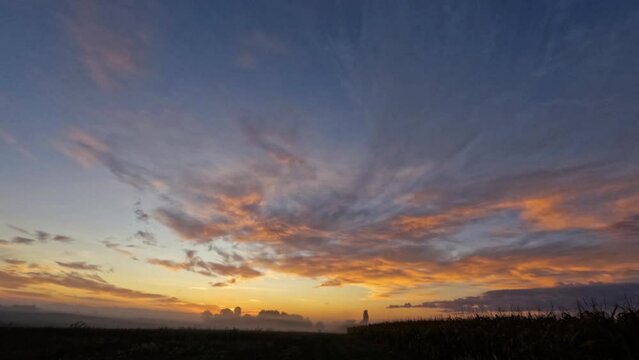 Beautiful time-lapse sunrise over a field between trees in a valley. Red and purple clouds run across the clear sky. A light fog hangs over the valley
