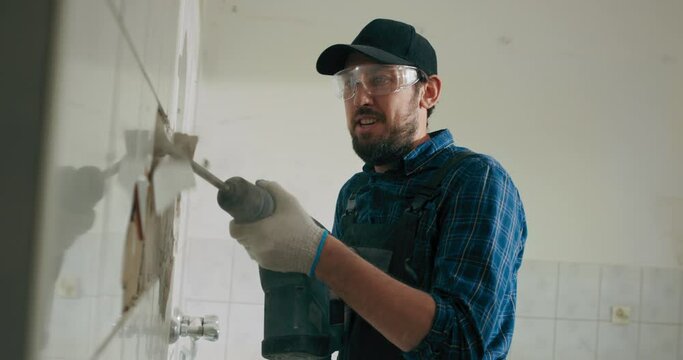 A Man In Work Clothes Wearing A Shirt And Cap Is Hammering Tiles In The Kitchen Bathroom Doing Renovations In The House.