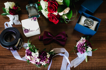 Groom's accessories: watch, perfume, tie-bow, belt, flowers on a wooden brown background. Top view.