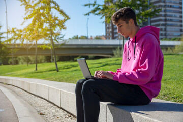 Young man sits on step, using laptop and looks on his screen. Young man is blogging, chatting online,checking email. Student learning online. Sunny day. Lifestyle.