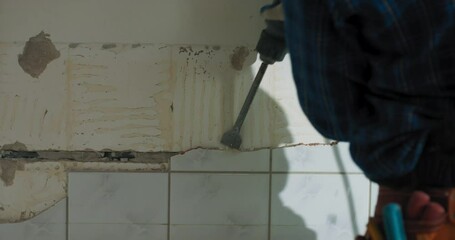 A man in work clothes wearing a shirt and with a tool belt is hammering tiles with a hammer in the kitchen bathroom doing renovations in the house.