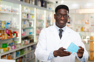 Male pharmacist in gown demonstrating drug package in salesroom of drugstore