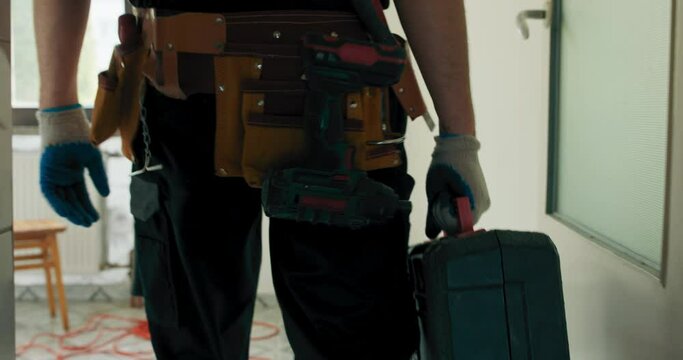 A man in work clothes with a toolbox prepares to renovate an apartment, chipping tiles, unpacks a suitcase with a hammer.