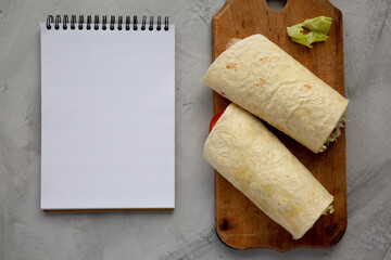 Homemade BLT tomato lettuce bacon Wrap with Mayo on a wooden board on a gray background, blank notepad, top view.