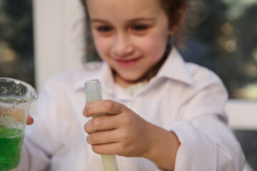 Details: Hands of Caucasian mischievous little girl in lab coat, holding a laboratory test tube and flask with chemicals and reagents, watching a taking place chemical reaction in the Chemistry class