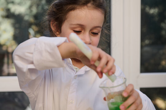 Focus on a mischievous little girl, a smart preschool child, in a white lab coat, conducting a chemical experiment, pouring liquid from test tube into a lab flask, learning Chemistry in science class