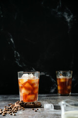 Iced coffee in a tall glass with poured cream, ice cubes and grains on an old rustic wooden table. Cold summer drink with pipes on black background with copy space 