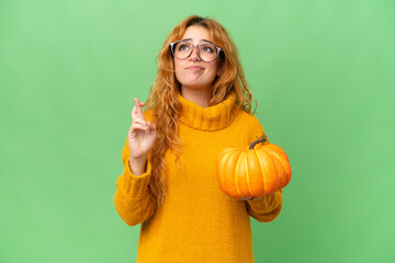 Young caucasian woman holding a pumpkin isolated on green screen chroma key background with fingers crossing and wishing the best - Powered by Adobe