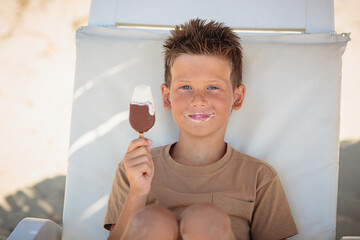 Handsome boy sitting on the beach wearing a t-shirt and eating ice cream on a stick