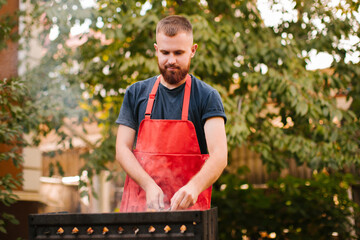 A young man in a blue t-shirt and a red apron with a beard is preparing a barbecue on the grill in...