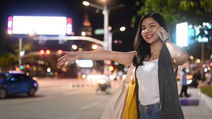 Fototapeta premium Smiling asian woman talking on phone and hailing for taxi on city street with road and buildings in background