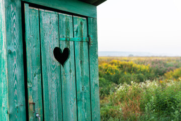 Heart shape on the old wooden toilet door, green toilet in the field. Outdoor recreation. Close-up.