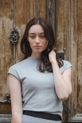 Beautiful portrait of Ukrainian girl with red hair, blue eyes and freckles in a grey summer dress posing with an old peeling brown wooden door in the background, vertical