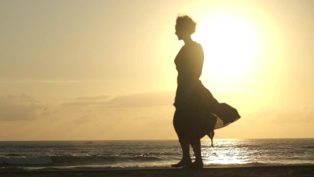 A young woman slowly moves with a graceful gait along the seashore. In the evening sunset, her silhouette looks especially attractive, mysterious and romantic.