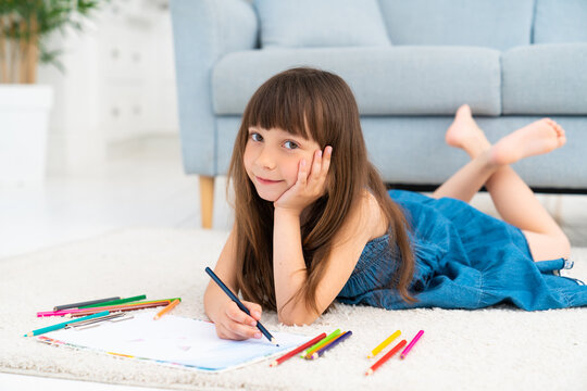 Cute funny girl in a denim sundress and bangs in her hair lies in the room on the carpet on the warm floor and draws with colored pencils. Homeschooling and creative development of the child.