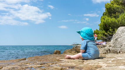 A baby with sunscreen clothes looks at the sea on vacation