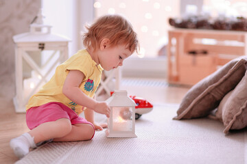 A charming two-year-old girl in bright clothes examines a burning white flashlight while sitting on the floor. The concept of familiarizing the child with the environment in the house